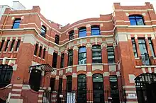 Façade de l'école en briques. La porte d'entrée se situe en haut d'un petit escalier, derrière un portail en fer forgé.