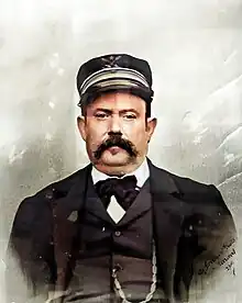 Photo en noir et blanc en buste d'un homme en costume portant une casquette et une moustache.