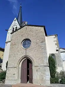 On voit la façade d'une église. À gauche se dresse un clocher surmonté d'une flèche. Une rampe facilite l'accès à la porte d'entrée notamment pour les usagers de fauteuils roulants. Au dessus de l'entrée se trouve un vitrail circulaire tandis qu'à droite, derrière un arbre se cache un autre vitrail.