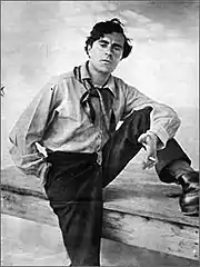 Photo noir et blanc d'un homme en manches de chemise, postérieur et pied gauche sur une table, foulard au cou, cigarette à la main