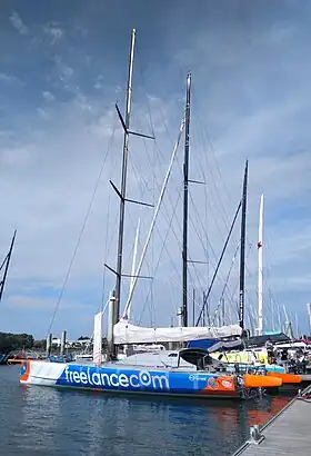 Vu de trois quarts arrière en entier, voilier rouge, blanc et bleu amarré au ponton. Pont et rouf gris.