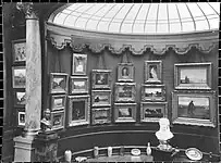Vue de la rotonde numéro 1 de la Galerie Vasnier avec la verrière comme source de lumière zénithale. Le mur est recouvert de tableaux, devant sont disposés deux bustes sur leur socle et des objets d'art à même le sol.