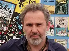 Photographie d'un homme posant devant une affiche reprenant des couvertures de livres.