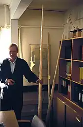 Un homme en costume se tient debout à gauche de la photo, tenant une longue dent de narval torsadée de la main droite. L'objet dépasse très largement la taille de l'homme et atteint presque le plafond.