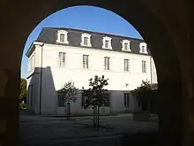Photo prise sous un proche dont l'arrondi sombre coupe le ciel bleu ; on voit un bâtiment de deux étages aux murs blancs, le toit mansardé est fait d'ardoises.