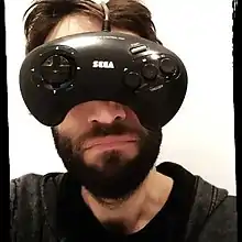 Portrait d'un homme brun barbu, qui met une manette de jeu vidéo devant son visage comme si c'était une paire de lunettes.