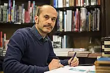 un homme dégarni avec une moustache en train de dédicacer dans une librairie.