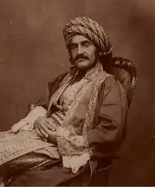 Photographie sépia d'un homme vêtu d'un habit du Moyen-Orient du XIXe&nbsp;siècle, portant une large moustache, assis sur une chaise avec les mains croisées sur ses jambes