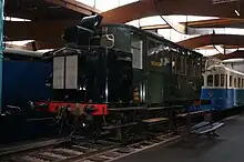 Vue de trois quart avant d'un petit autorail de forme parallélépipédique en livrée vert sombre.