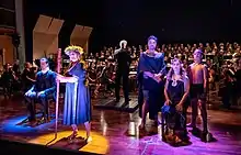 Sur le devant de la scène, les 5 comédiens de l'opéra en costume ; en arrière-plan, l'orchestre.