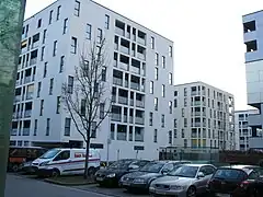 Vue de quelques bâtiments blancs identiques et carrés, ornés de nombreuses fenêtres rectangulaires, et d'un parking à l'avant-plan.