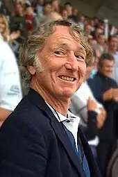 Photo en buste d'un homme en costume bleu se tenant de trois-quart dans une tribune de stade.