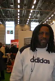 Portrait d'un homme. Coiffé de dreadlocks, il porte un pull à dominante blanche ayant l'inscription de sa marque, allemande, écrite en noir.