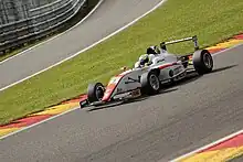 Photographie d'une monoplace de F4 blanche et rouge, vue de trois-quarts gauche, sur une piste sèche, montant le virage pentu du Raidillon.