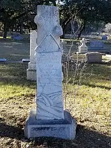 stèle de tombe, sculptée en forme de tronc d'arbre étêté auquel est accroché une bannière portant l'inscription tombale.