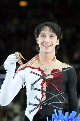 Homme souriant sur un podium et mettant en avant sa médaille. Il porte un costume bicolore blanc et noir.