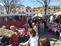 Le marché des forains fait partie de l'identité de la place - Des gens font leurs courses, marchent, discutent, observent des habits. La scène se déroule entre plusieurs stands de vêtements et de fripes. On aperçoit un stand de fruits et de légumes à l'arrière plan.