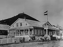 Photographie en noir et blanc d'une maison avec un étage qui fait flotter le pavillon de l'Empire russe. La maison, en bois, est sur le bord d'une rue assez large, et au fond se distingue une montagne.