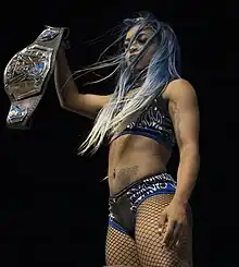 Une catcheuse aux cheveux bleu brandissant dans la main droite une ceinture de championne de catch.