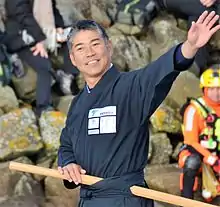 Dans le chenal des Sables-d'Olonne, en buste, souriant, il salue la foule. Il est vêtu d'un kimono gris sombre et porte un sabre de bois à la ceinture.