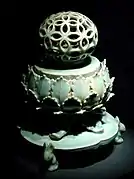 Photographie d'un objet constituée de trois parties. Un socle dont les pieds sont en forme de lapin, un corps central en forme de fleur, et un sommet en forme de globe creux.