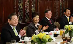 Photographie d'un banquet, au centre Park Geun-hye est en train d'applaudir entourée par trois hommes âgés.