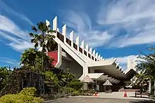 Photographie d'un grand bâtiment rouge et blanc en prisme triangulaire. Au premier plan se trouve un petit jardin composé de palmiers, de parasols, d'une cascade et l'inscription « Muzium Sabah ».