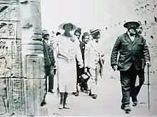 vêtue de clair, la reine chemine en compagnie du professeur Capart dans une allée d'un temple dont on aperçoit à gauche l'un des murs gravés de hiéroglyphes