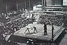 Photographie montrant deux hommes luttant et un juge sur un ring au milieu des spectateurs, depuis un point de vue en hauteur.