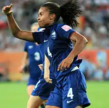 Photographie d'une joueuse de football noire, vue de profil, souriante, levant le bras droit.