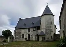 Photographie en couleurs d'un bâtiment à toit pentu accolé d'une galerie en hauteur et d'une tourelle octogonale, la façade d'un deuxième bâtiment visible sur la droite, un arbre s'élevant sur la gauche, une étendue herbeuse apparaissant au premier plan.