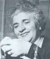 Image en noir et blanc d'un visage de femme, cheveux frisés blancs regardant avec un sourire un objet tenu dans sa main.