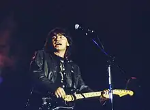 Photographie en couleur d'une personne sur scène, cheveux noirs, tenant une guitare et chantant, devant un haut micro sur pied