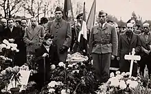 Photographie en noir et blanc d'une foule se recueillant dans un cimetière. Au premier plan, une femme accroupie au milieu d'un parterre de bouquets de fleursv, à côté d'une tombe sur laquelle on peut lire « Manoushian ».