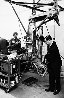 Photo noir et blanc, price dans un atelier. La jambe de train est totalement démontée, un technicien l'inspecte.