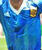 Photographie en couleurs. Gros plan sur l'écusson contrefait de la fédération argentine de football cousu sur un maillot bleu plus foncé.