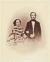 À gauche Marie-Henriette, les cheveux foncés divisés par une raie et portant une robe à crinolines et à passants horizontaux, marquant la taille, tient le bras droit de Léopold, revêtu d'une longue redingote sombre, portant une cravate sombre enserrant un col clair, son visage est recouvert de longs favoris foncés