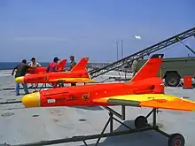 Trois drones rouges fuselés avec rampe sur pont de porte-avion. Les ailes delta et les nez sont jaunes (3&nbsp;×&nbsp;2,5&nbsp;m env.).