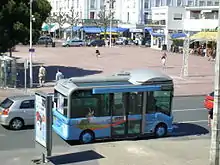 Photographie d'un minibus rectangulaire roulant le long de la place principale. Le véhicule surbaissé présente de grandes baies vitrées teintées de vert, un éclairage zénithal, de larges portes de verre en son centre et une carrosserie peinte en bleu vif décorée d'un paysage touristique et de divers logos de la région