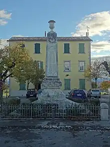 Le monument est un obélisque sur socle, surmonté d'une urne funéraire, et entouré d'une grille. Au fond, la façade de la mairie jaune un peu cachée par un arbre.