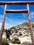 Photo couleur : vue en gros plan d'un torii, portique d'entrée d'un sanctuaire shintō, au sommet d'une montagne au sol parsemé de rochers, sous un ciel bleu nuageux.