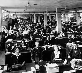 Une grande salles où des secrétaires tantôt en uniforme, tantôt en civile, travaillent devant des machines à écrire. Au premier plan, deux sous-officiers supervisent le service.