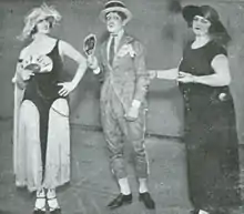 Photographie en noir et blanc. Trois personnes se tiennent debout et ont un chapeau sur la tête : un homme est au milieu et à gauche et à droite se trouvent deux femmes. Les deux femmes portent une robe. La robe à gauche de l'image et bicolore blanc/noir, la robe à droite est noire. L'homme est vêtu d'un complet gris.