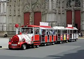 Photographie en couleurs d’un petit train routier touristique, composé d’une locomotive ressemblant à une locomotive à vapeur et de trois wagons sans vitres, le tout rouge et blanc, dans une rue devant un édifice religieux.