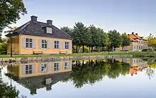  Bâtiment sans étage d'allure classique au bord d'un étang rectiligne. Quelques bancs, collés au bâtiment, au bord de l'étang.
