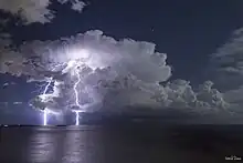 Un éclair, en arrière-plan, descend d'un nuage pour toucher la mer, au premier plan, et illumine le ciel de l'aube