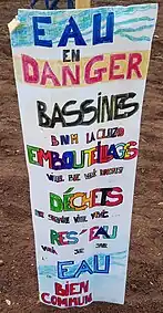 Une pancarte en bois blanche, plantée dans un champ labouré, sur laquelle sont écrits en couleur différents slogans anti-bassines