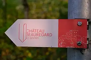 Panneau séparé en deux couleurs : à droite du rose et à gauche du blanc. Sur la partie blanche se trouve le nom du château avec la notion « le sentier » et sur la partie rose se trouve un dessin du château comme il devait être au Moyen Âge : plusieurs blocs aux sommets crénelés.