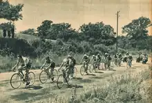 Photo en noir et blanc d'un peloton cycliste sur une route de campagne bordée de champs.