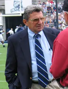 Un homme âgé en costume, mains dans les poches, portant des lunettes teintées, sur un terrain de football américain.
